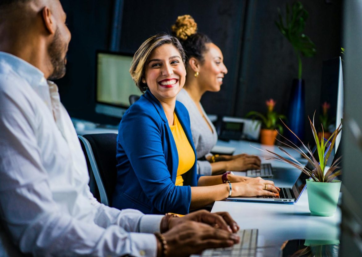 Three diverse professionals working and smiling at office desks, fostering teamwork and collaboration. Töö