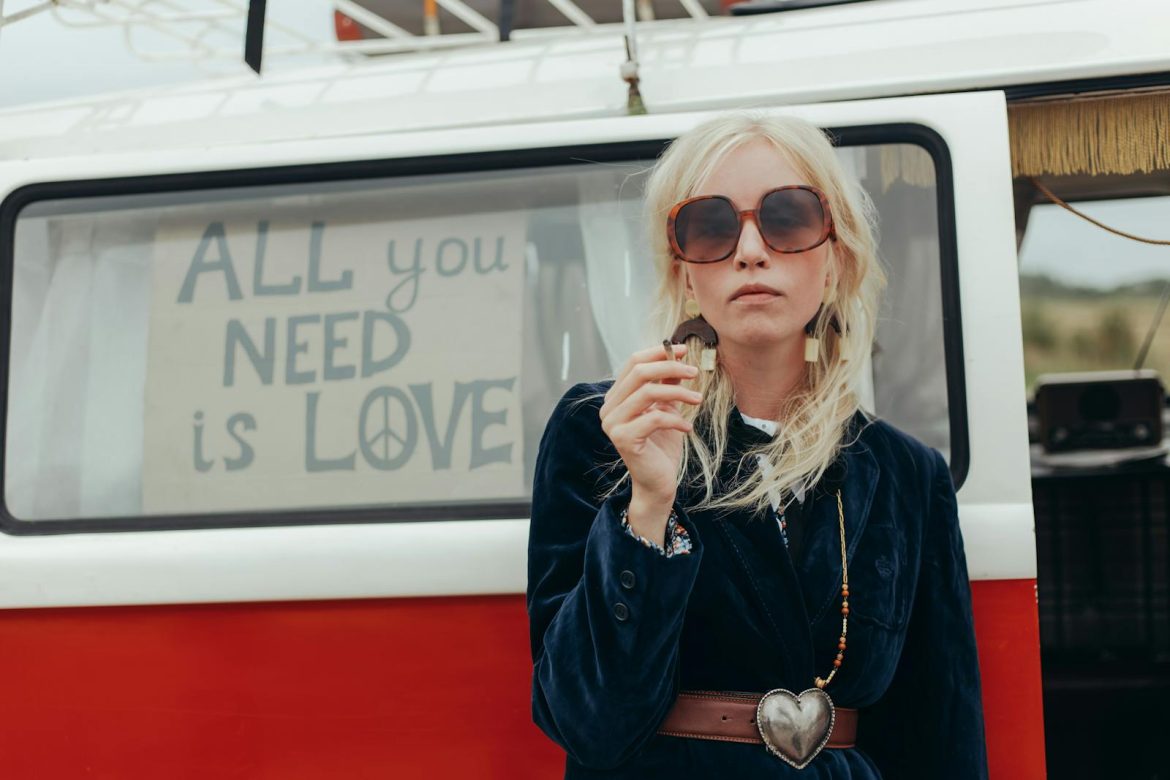 Fashionable woman in front of a retro van with 'All you need is love' sign, capturing the hippie vibe.