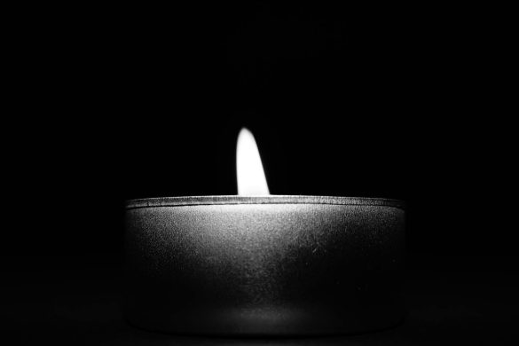 Black and white close-up of a single tealight candle, emitting soft light in the dark.