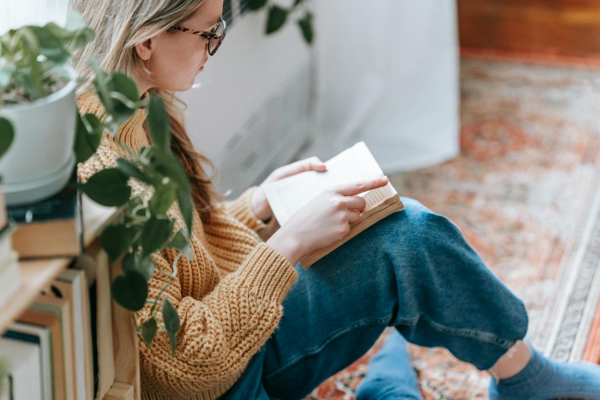 Side view of crop female student in casual clothes sitting on carpet near bookshelf with plant and reading book at home