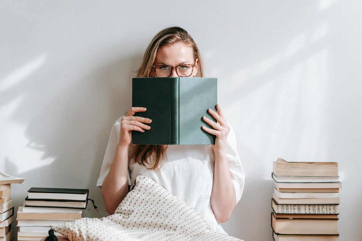 Young woman with eyeglasses reading book indoors, surrounded by books. Calm, thoughtful atmosphere.