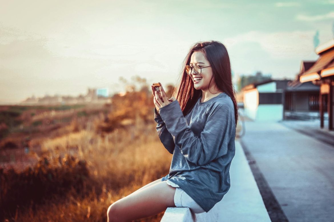 Happy woman with smartphone outdoors during sunset, capturing a joyful moment.