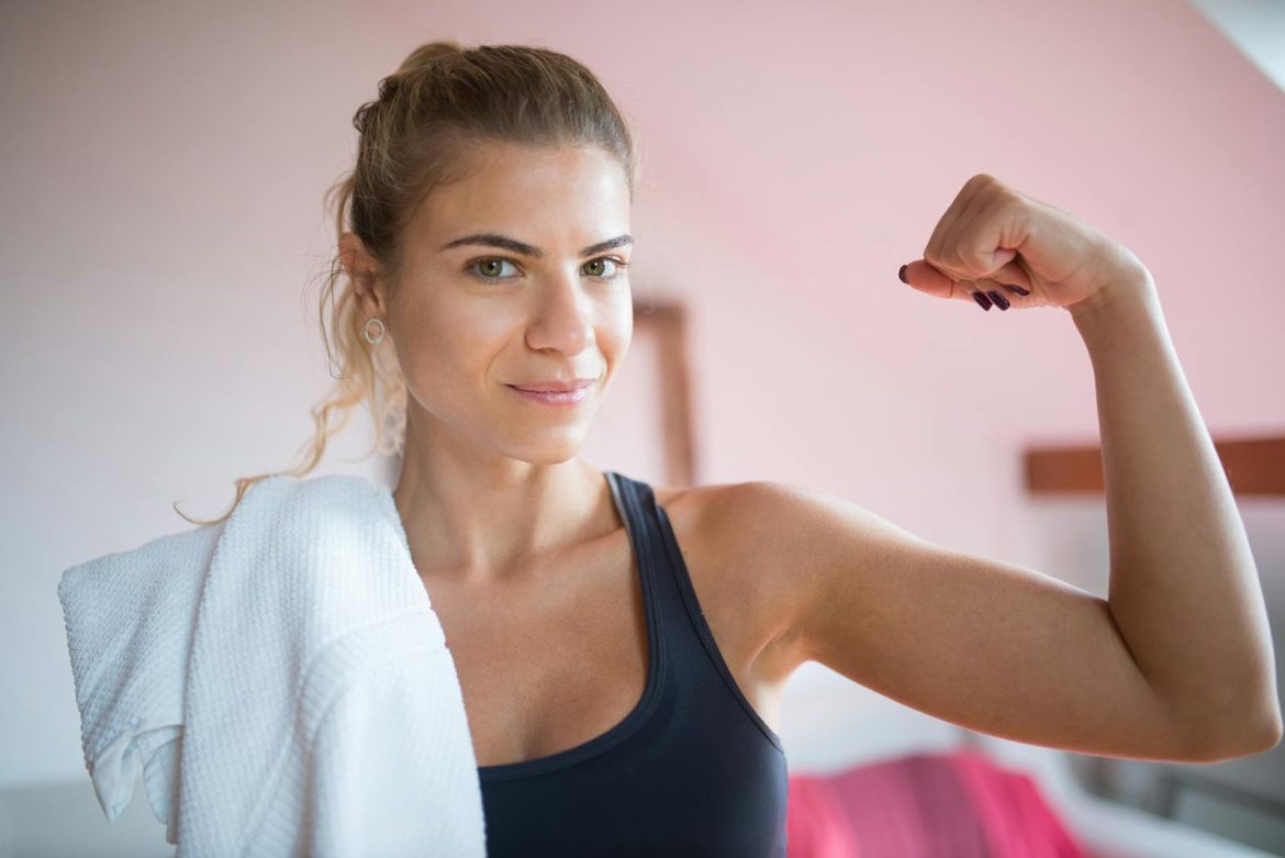 Confident woman in a tank top flexing her arm with a towel over her shoulder indoors.