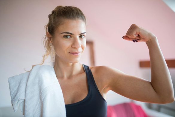 Confident woman in a tank top flexing her arm with a towel over her shoulder indoors.
