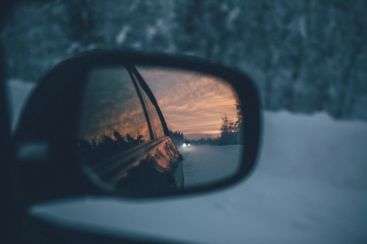 A scenic winter drive captured through a car's side mirror during sunset, reflecting snowy roads and trees.
