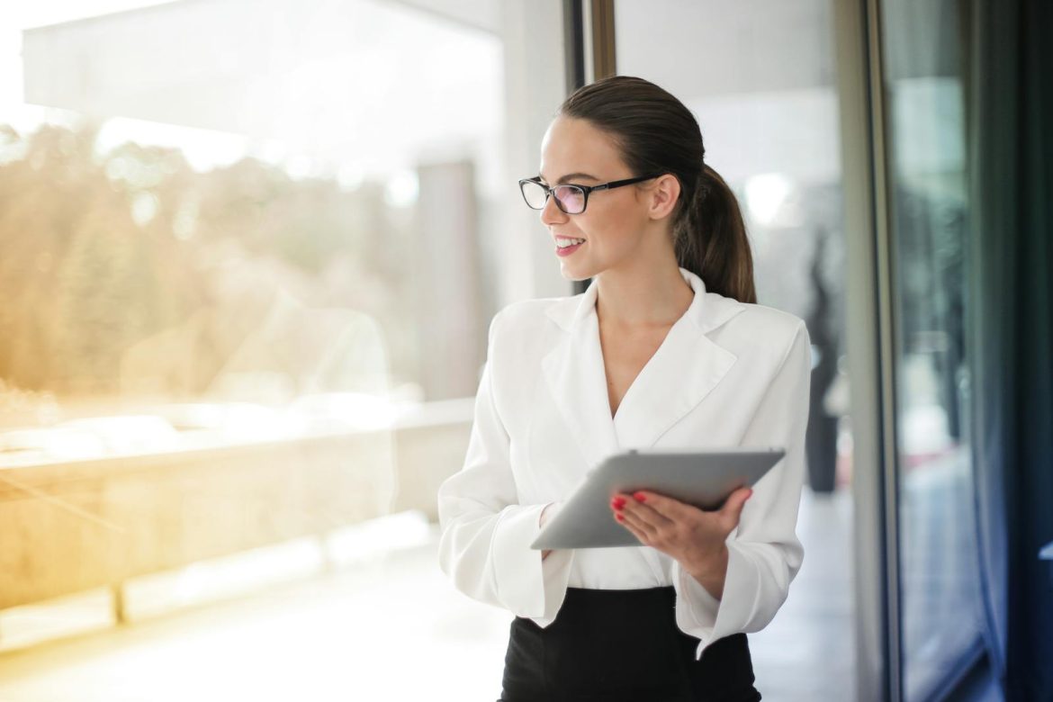 A professional woman in glasses holding a tablet in a modern office setting, exuding confidence.