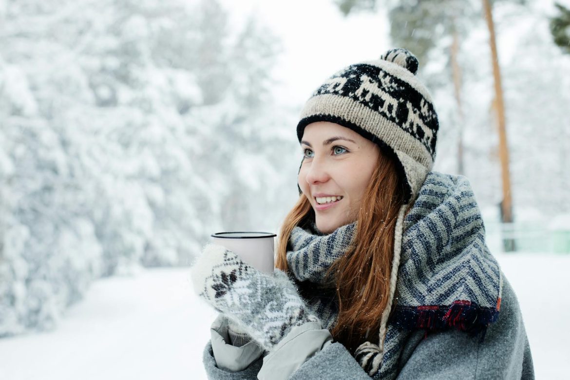 A woman in winter attire enjoying a hot drink outdoors in a snowy landscape, smiling happily.