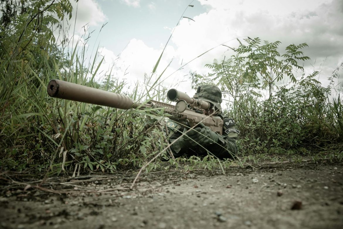 A camouflaged sniper with rifle hidden in grasslands during daytime surveillance.