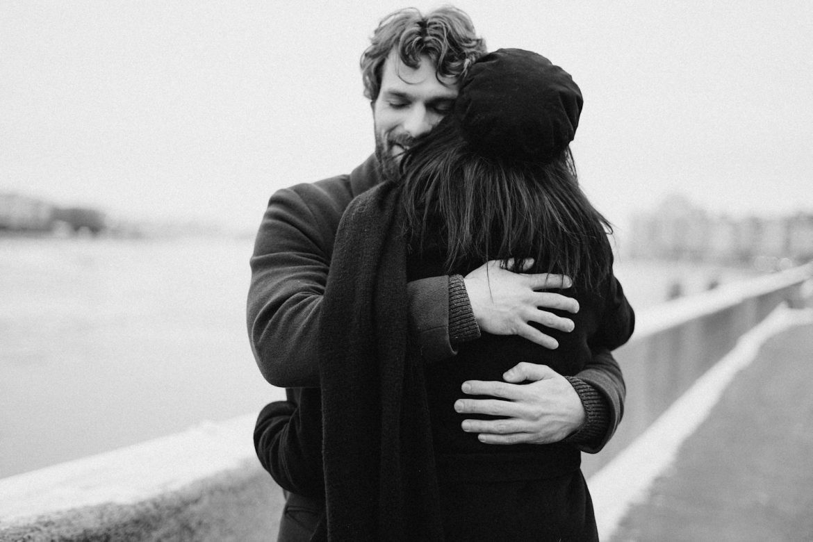 A romantic couple embraces warmly by the water on a winter day.