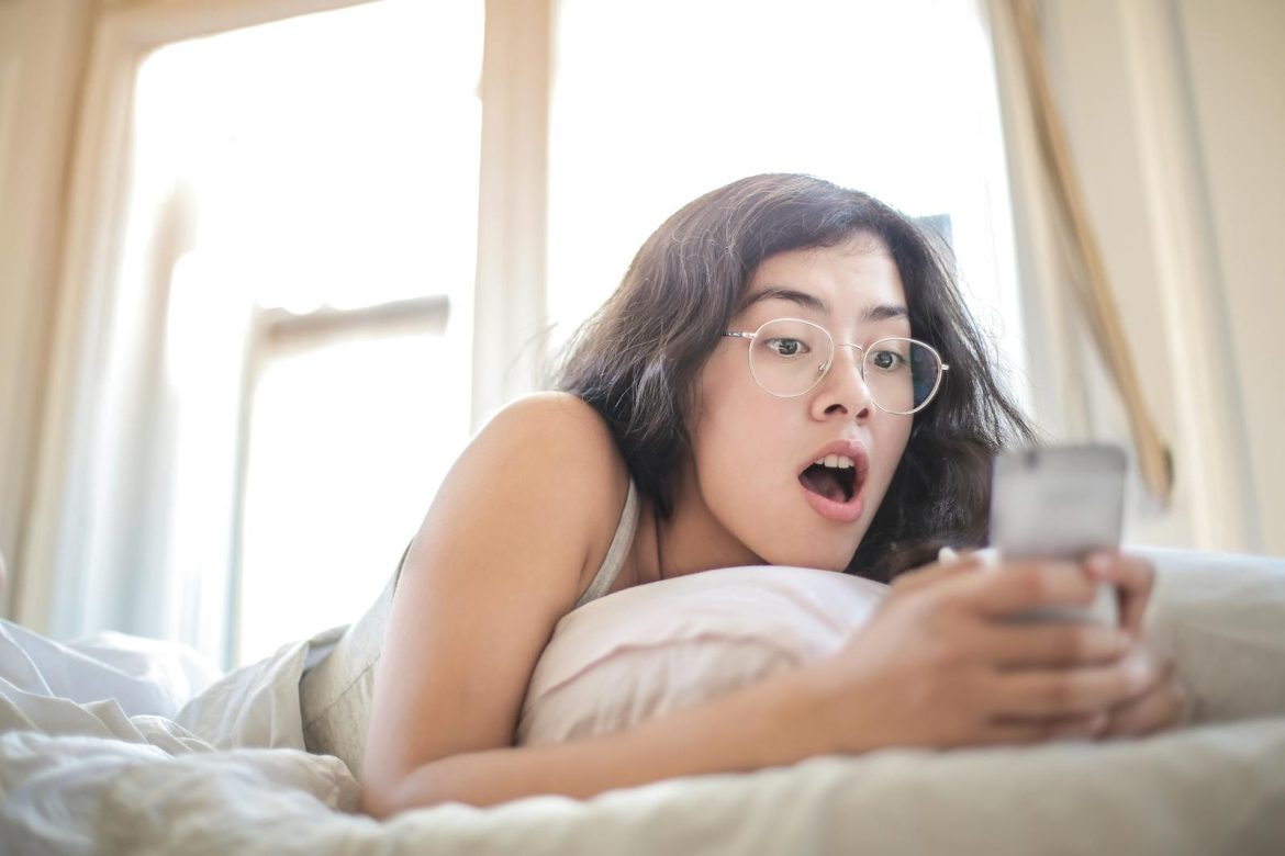 Young woman lying on bed, surprised by something on her smartphone, in a bright bedroom setting.