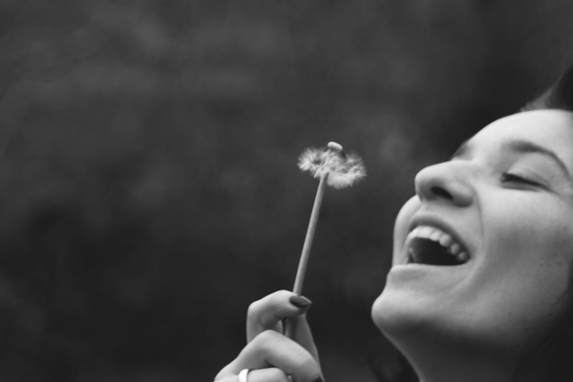 A joyful woman smiling while blowing on a dandelion in a black and white portrait.