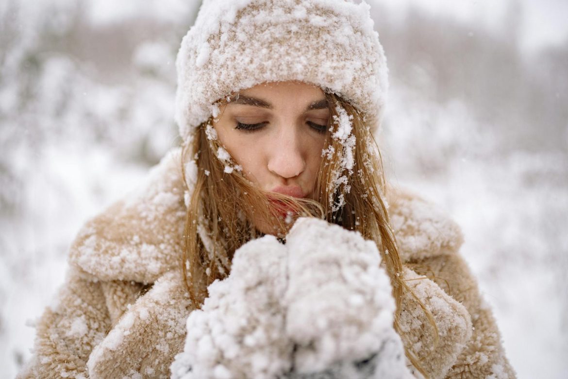 Close-up of a woman in winter attire surrounded by snow, blowing warm air into her hands.