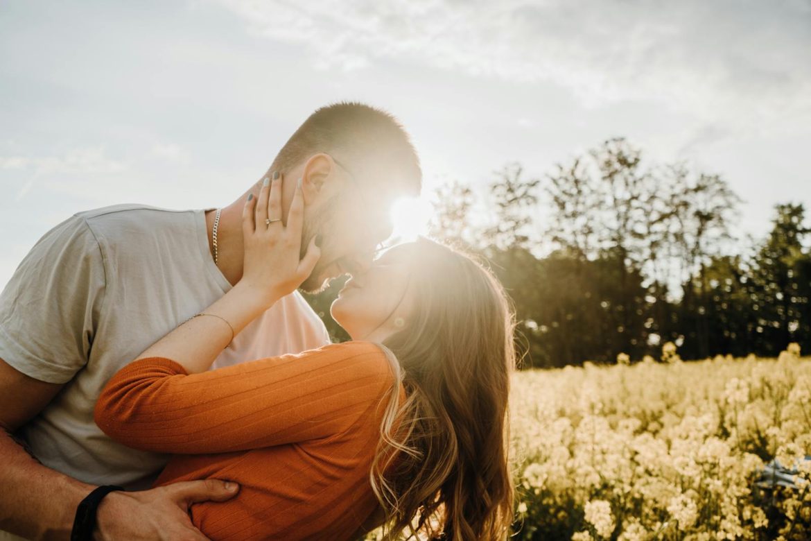 A couple shares a romantic embrace in a sunlit field with flowers and trees.
