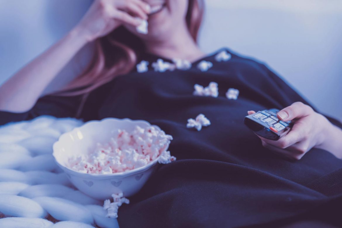 Woman relaxing on couch eating popcorn while watching TV, holding remote control, indoors.