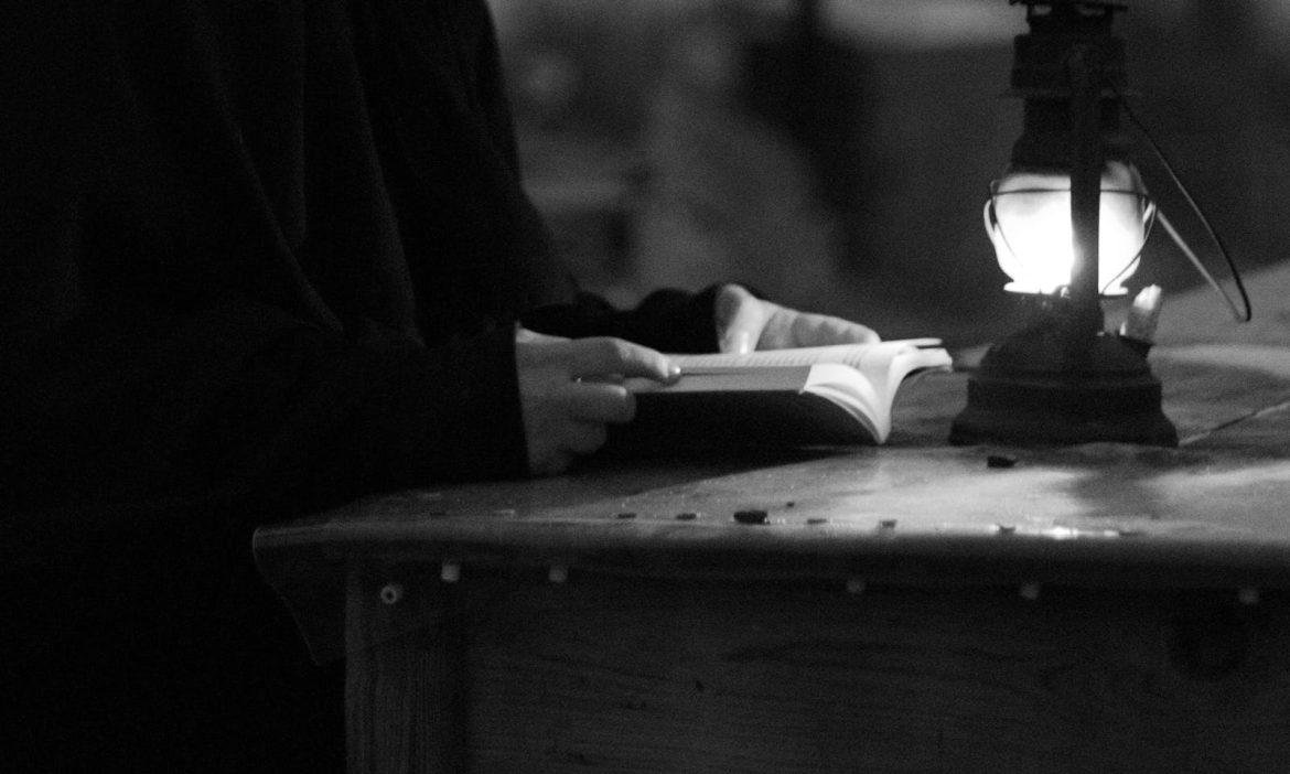 A person reads a book by an oil lamp, captured in a moody black and white photograph.