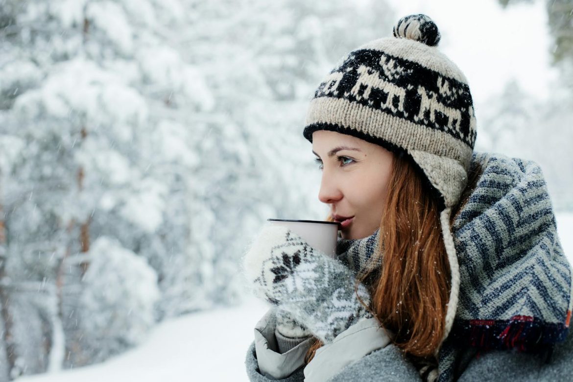 Woman in warm winter clothes enjoys a hot drink in snowy outdoor setting.