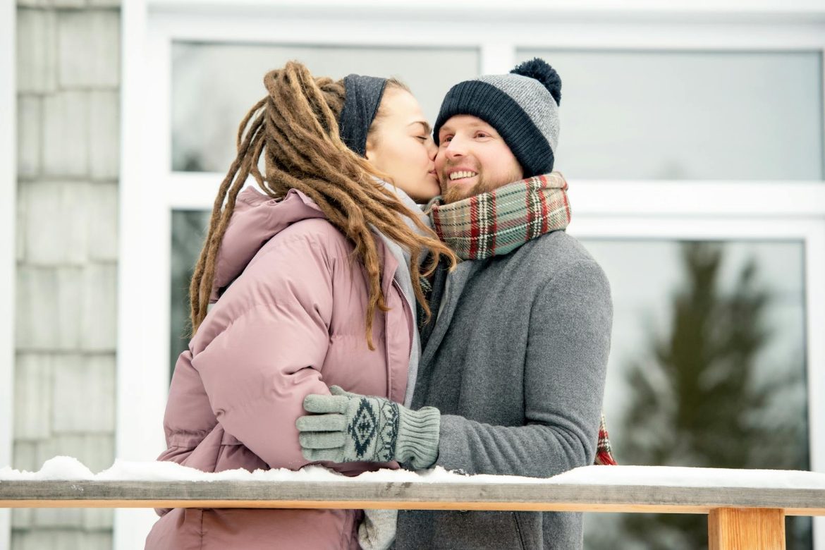A couple shares a warm kiss outdoors in a cozy winter setting.