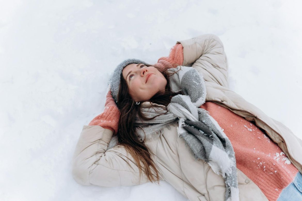 A happy woman lies in the snow, wearing cozy winter clothing and smiling as she looks up.