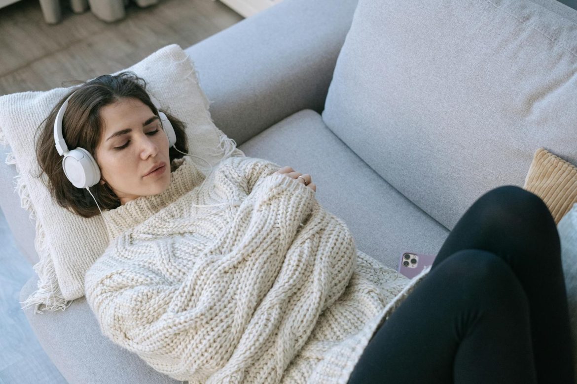 A woman in a cozy sweater relaxes with headphones on a comfortable couch indoors.