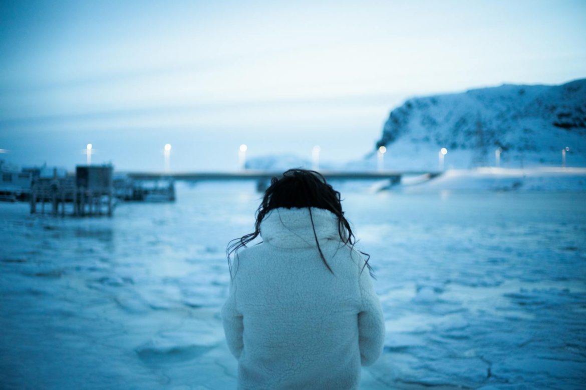 Woman in white fur coat overlooking frozen lake at dusk, embracing winter's beauty.