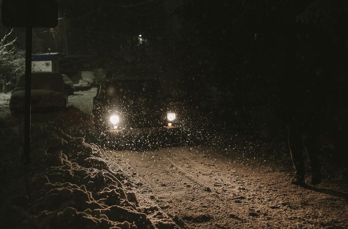 A car drives through a snow-covered road at night, with headlights illuminating falling snowflakes.