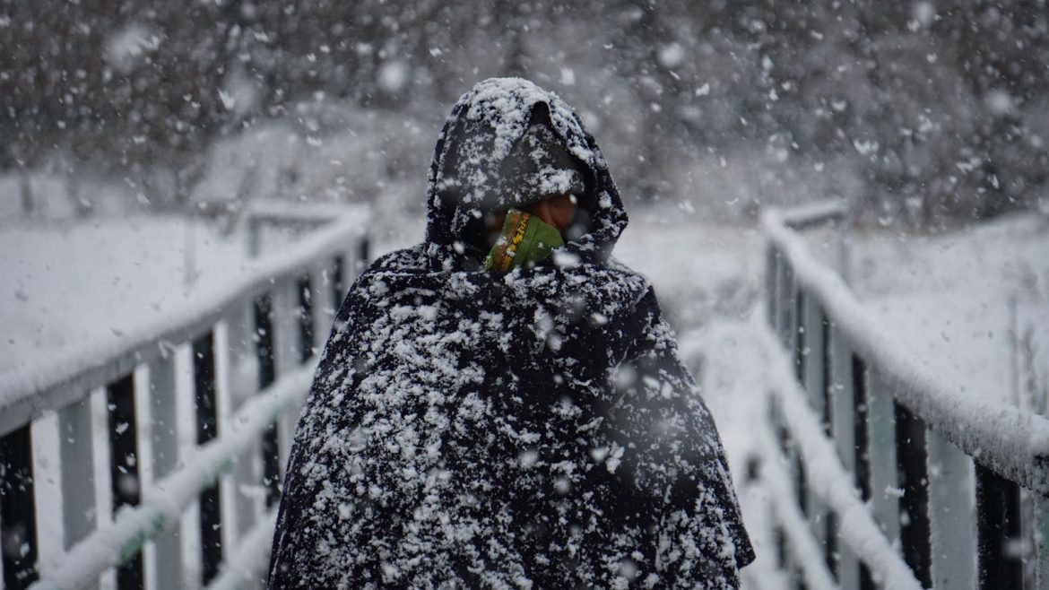 A woman stands on a snow-covered footbridge in Kashmir, wrapped in a black coat with heavy snowfall.