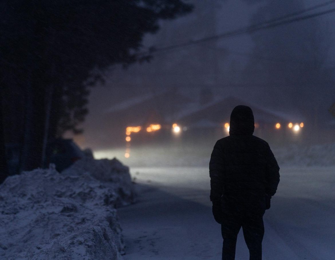 A solitary figure stands on a snow-covered road under dim, winter evening lights.