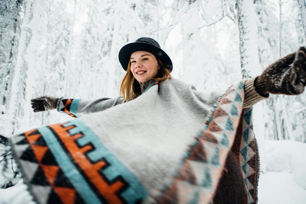 A cheerful woman wearing a geometric poncho and hat, enjoying a snowy winter forest scene.