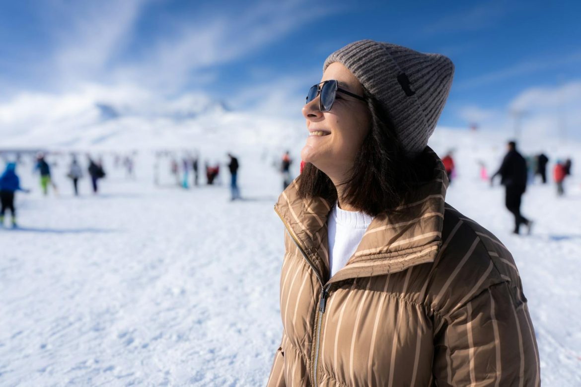Woman in winter attire smiling on snowy Mount Erciyes, Türkiye, under a bright sky.
