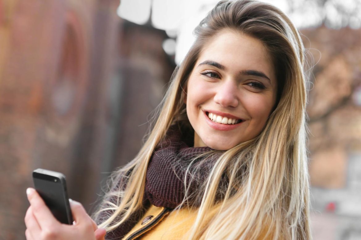 Young woman smiling while holding a smartphone, wearing a scarf and coat outdoors.