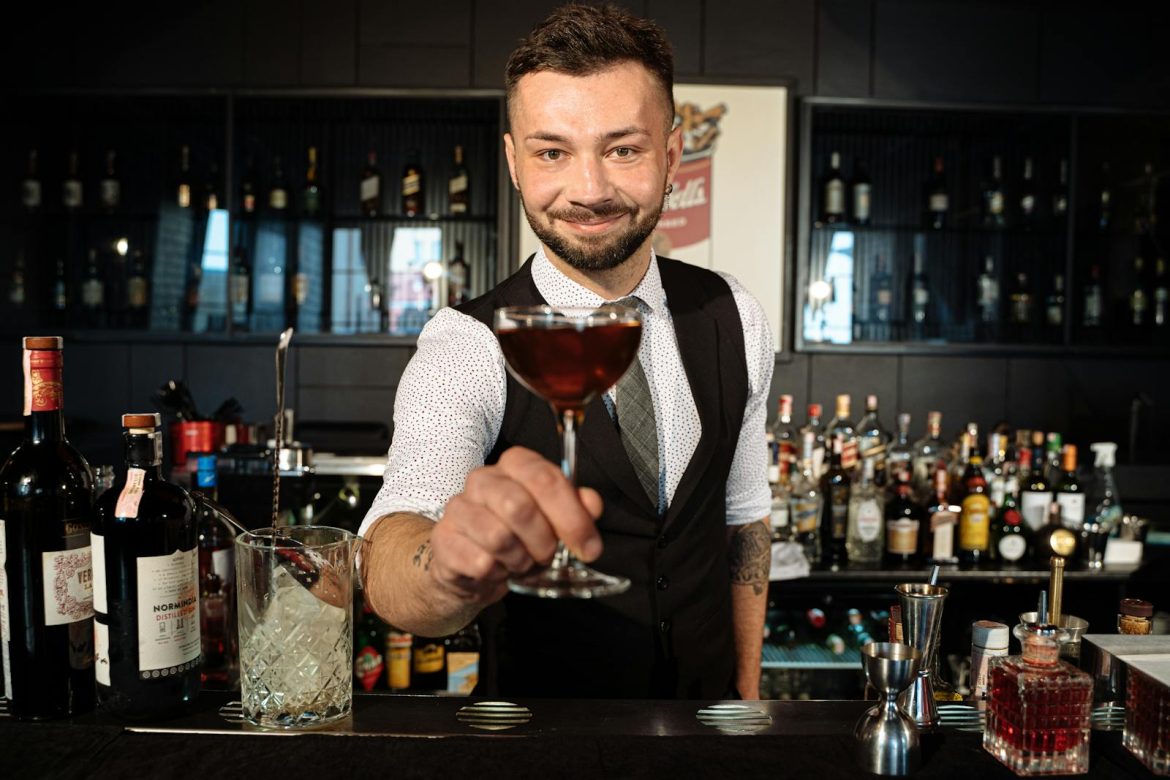 Smiling bartender in a black vest presenting a cocktail drink at a stylish bar with a range of bottles.