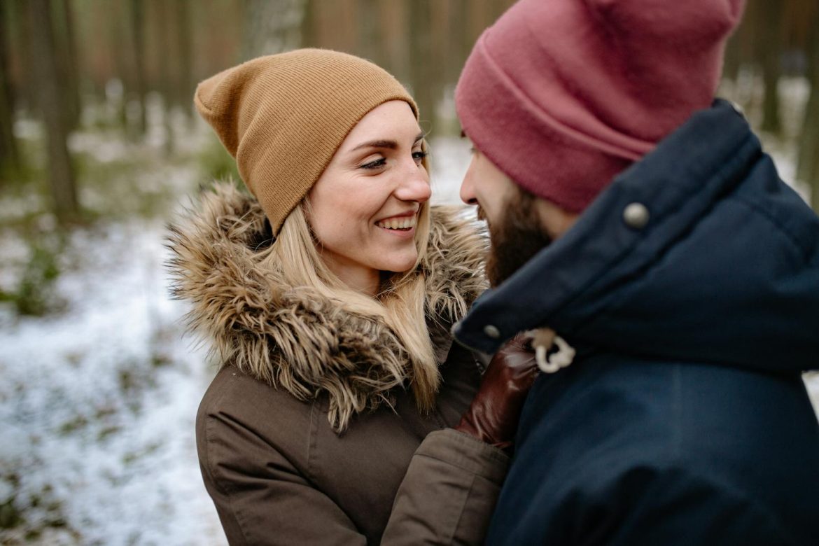 Happy couple embracing in a snowy forest, wearing winter outerwear and smiling lovingly.