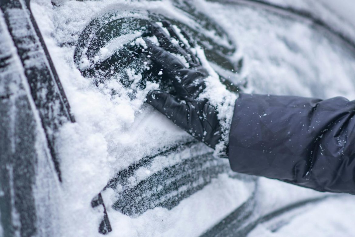 A close-up of a gloved hand clearing snow from a car window during winter.
