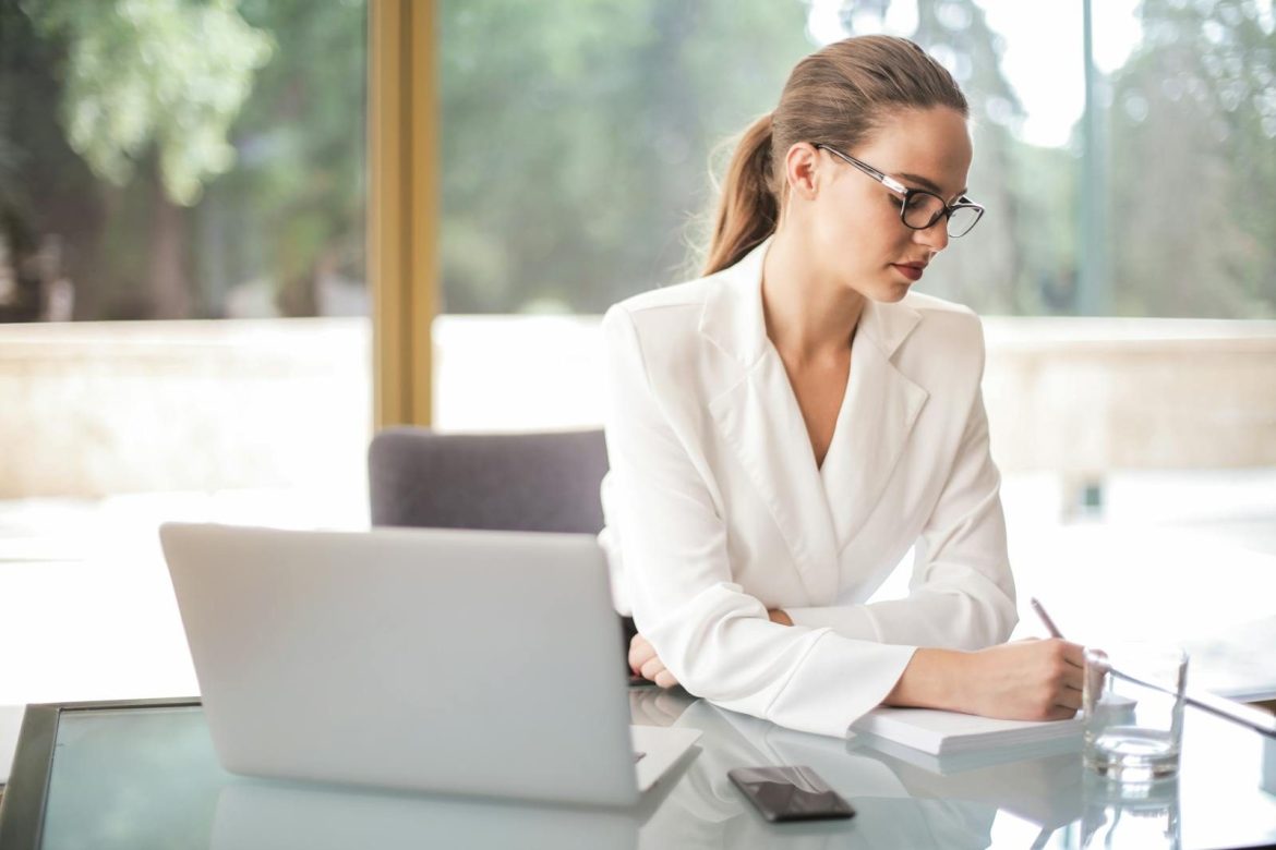 Pensive female entrepreneur in classy wear and eyeglasses sitting at table with laptop and writing information in planner while working on new project