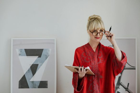 Woman in red robe holding a clipboard and pen, wearing eyeglasses indoors.