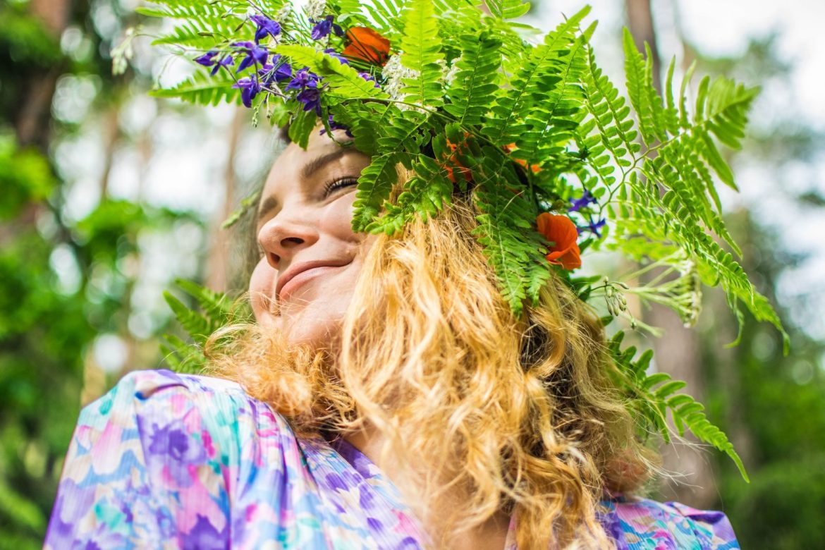 A joyful woman wearing a vibrant floral headdress, smiling in a lush outdoor setting.
