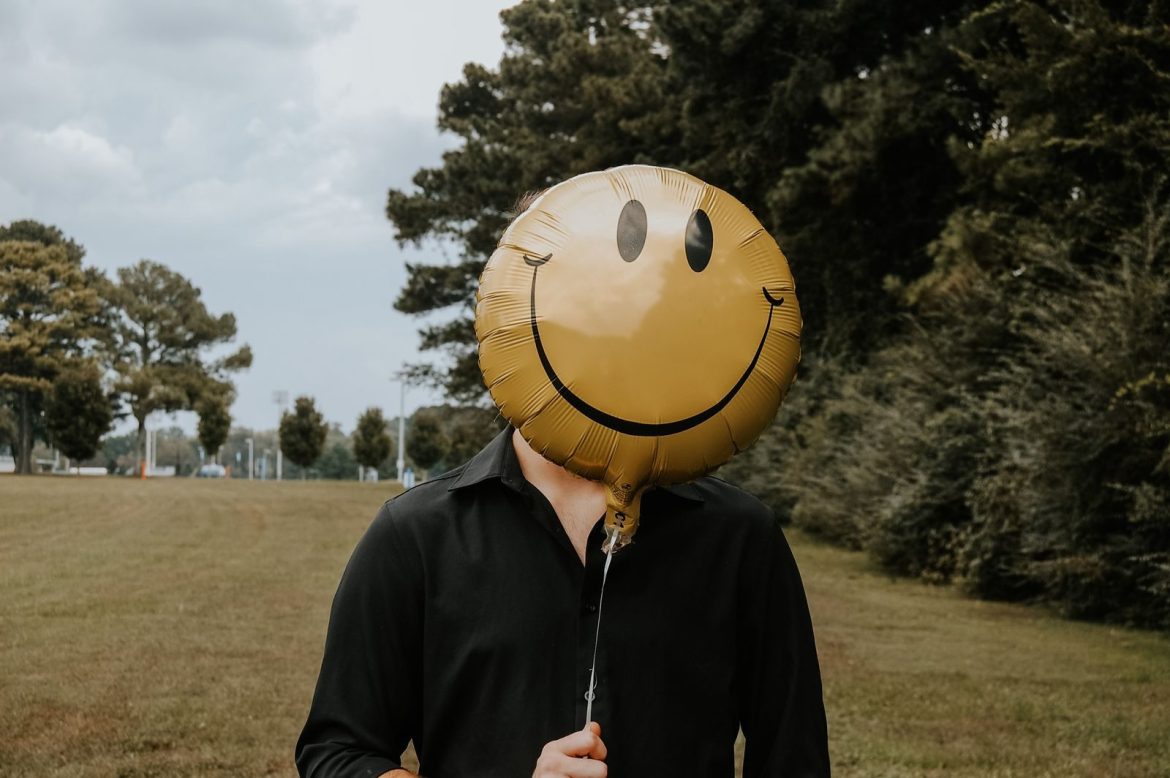 A person holds a smiley face balloon obscuring their face in an outdoor park setting.