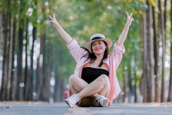 Smiling woman sitting cross-legged on a scenic tree-lined road, wearing a hat and raising peace signs.