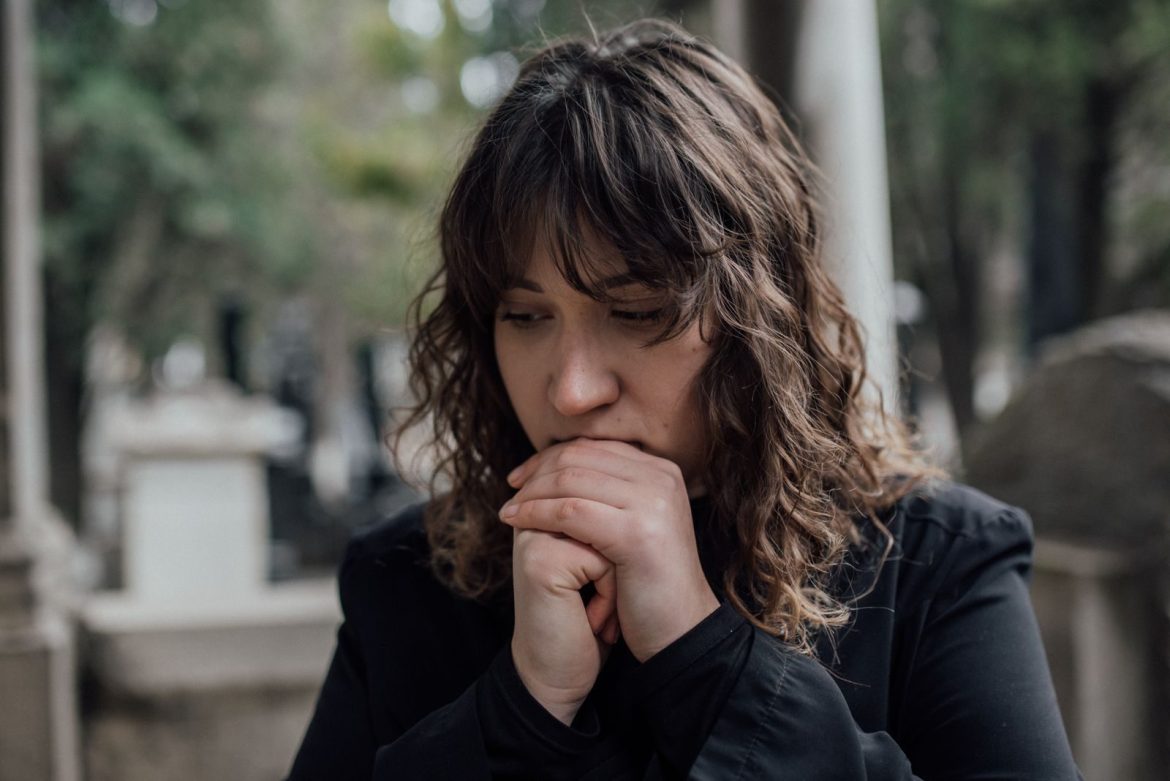 Pensive woman in black coat standing outdoors with hands together, conveying sadness.
