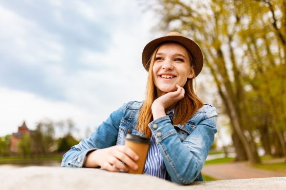 Casual woman in denim and hat enjoying a sunny day with coffee outdoors.
