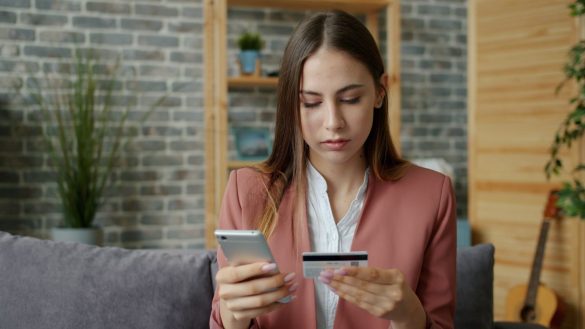 A young woman in a pink blazer shops online using a smartphone and credit card indoors.