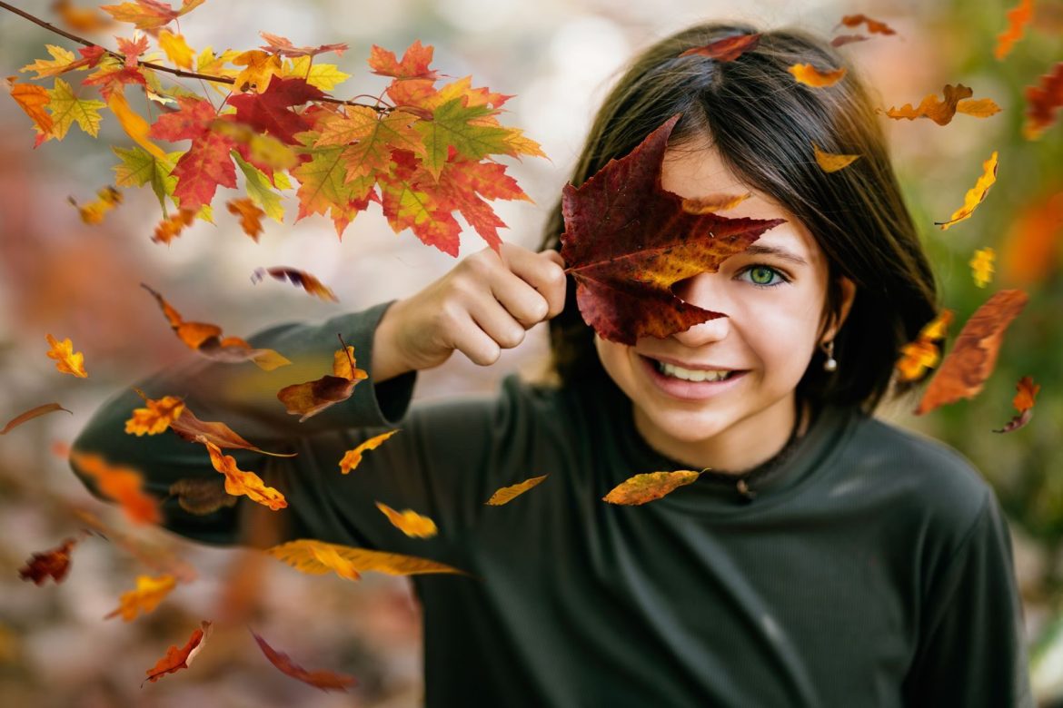 Content preteen girl with dark hair in casual clothes smiling while covering eye with tree dry leaf and looking at camera in autumn forest